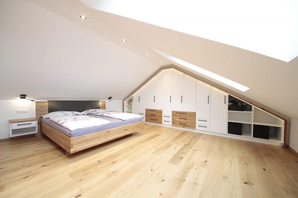 Großzügiges Dachgeschoss-Schlafzimmer mit schräger weißer Decke, Holzboden, Bett und eingebauten Stauschränken.