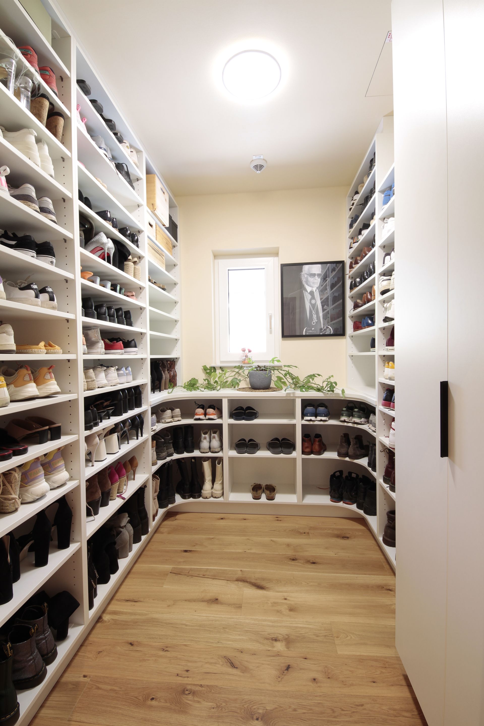 Schmaler begehbarer Kleiderschrank mit weißen Regalen, Schuhen an beiden Seiten und einem Holzboden unter einer Deckenleuchte