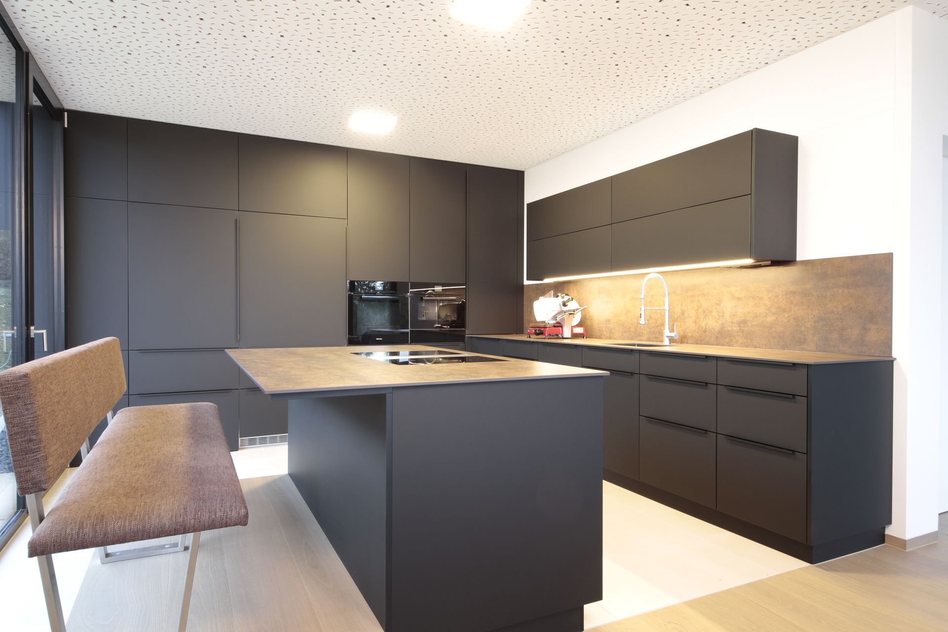 Moderne Küche mit mattschwarzen Schränken, Kochinsel, Holzrückwand und zwei gepolsterten Hockern