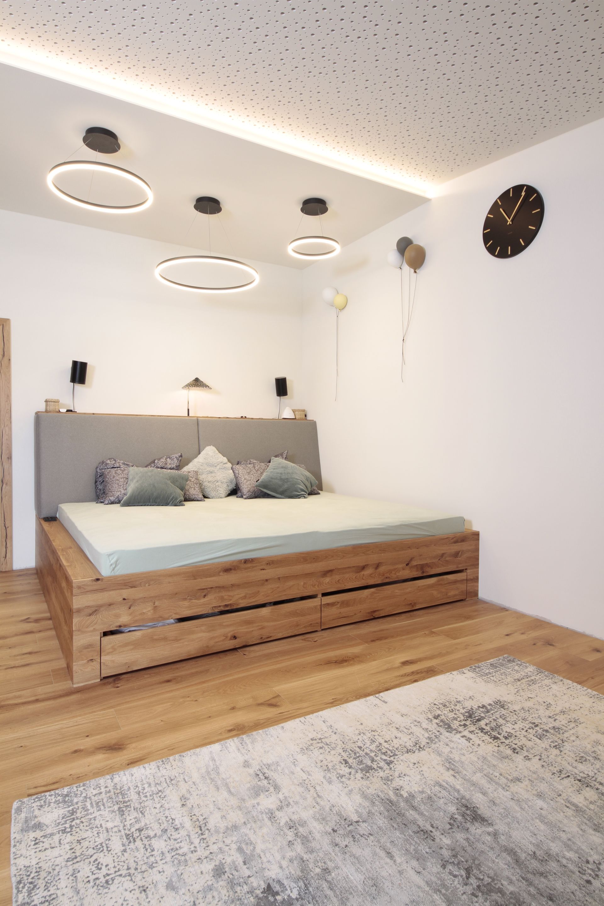 Minimalistisches, modernes Schlafzimmer mit Holzbett, grauem Kopfteil, runden Deckenleuchten und Wanduhr.