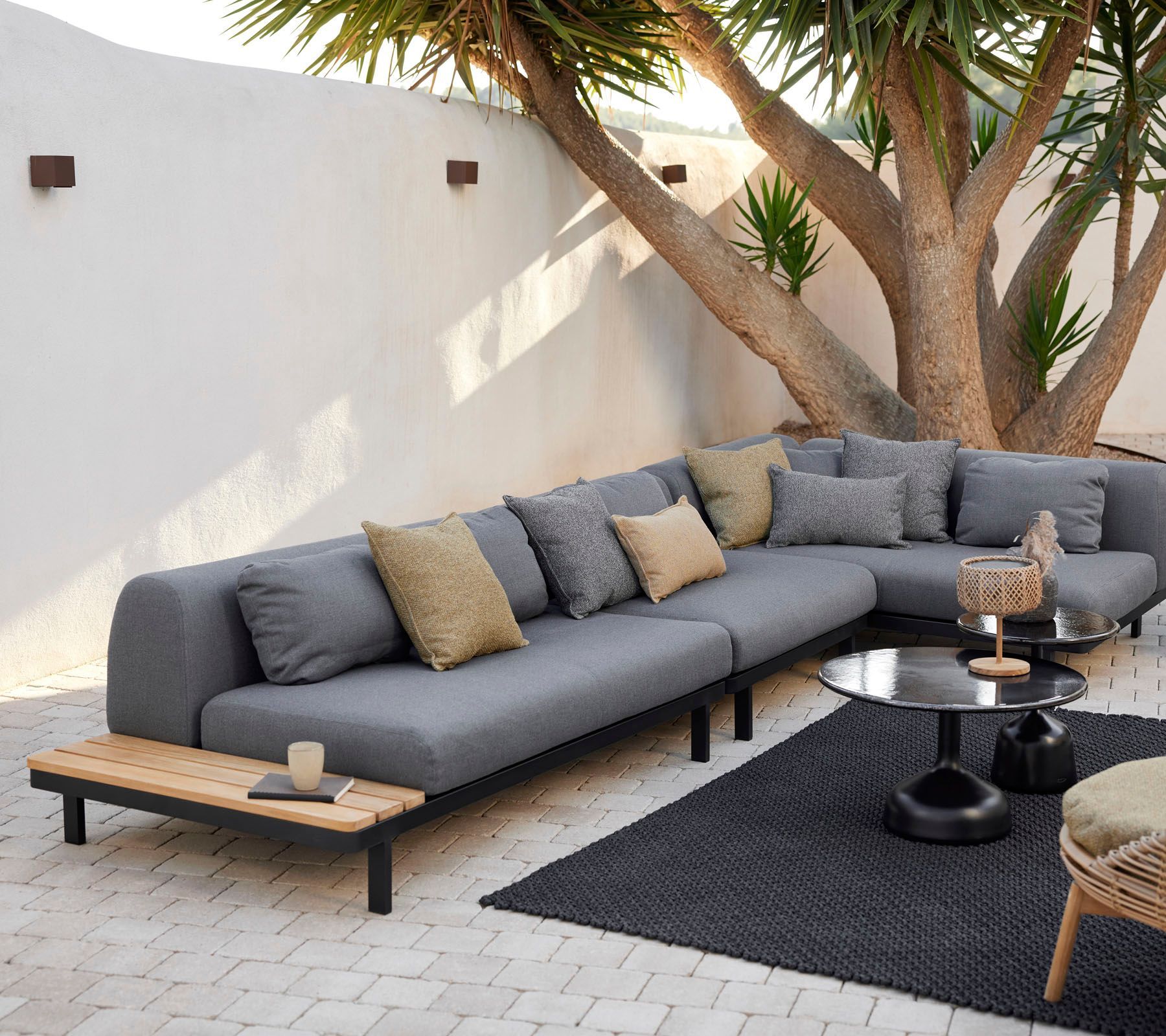 Moderne Außenterrasse mit grauem Ecksofa, Holzbänken, schwarzem Teppich und Bäumen vor einer weißen Wand
