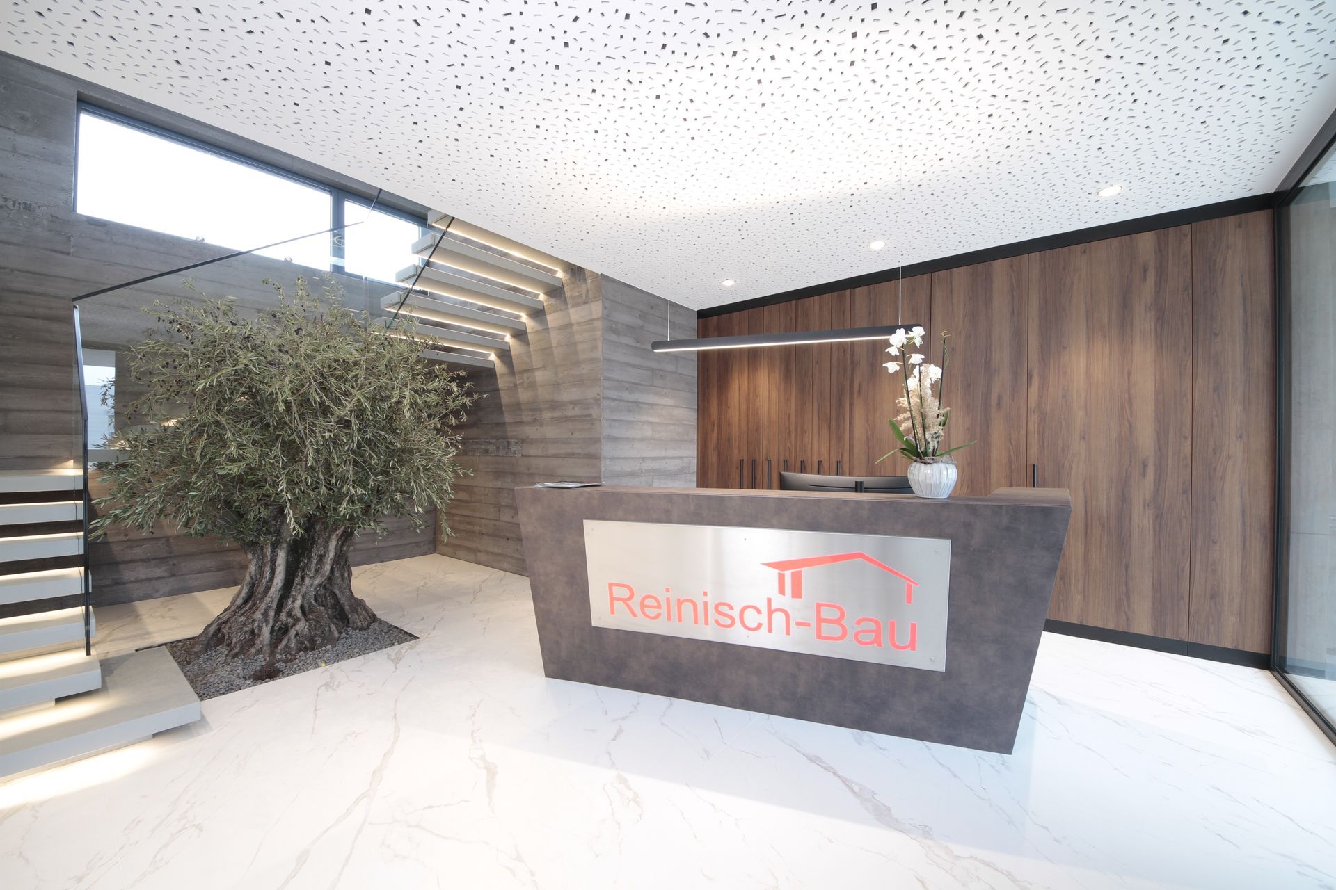 Moderne Bürolobby mit steinernem Empfangstresen, holzgetäfelten Wänden, Treppenhaus und großem Baum im Innenbereich