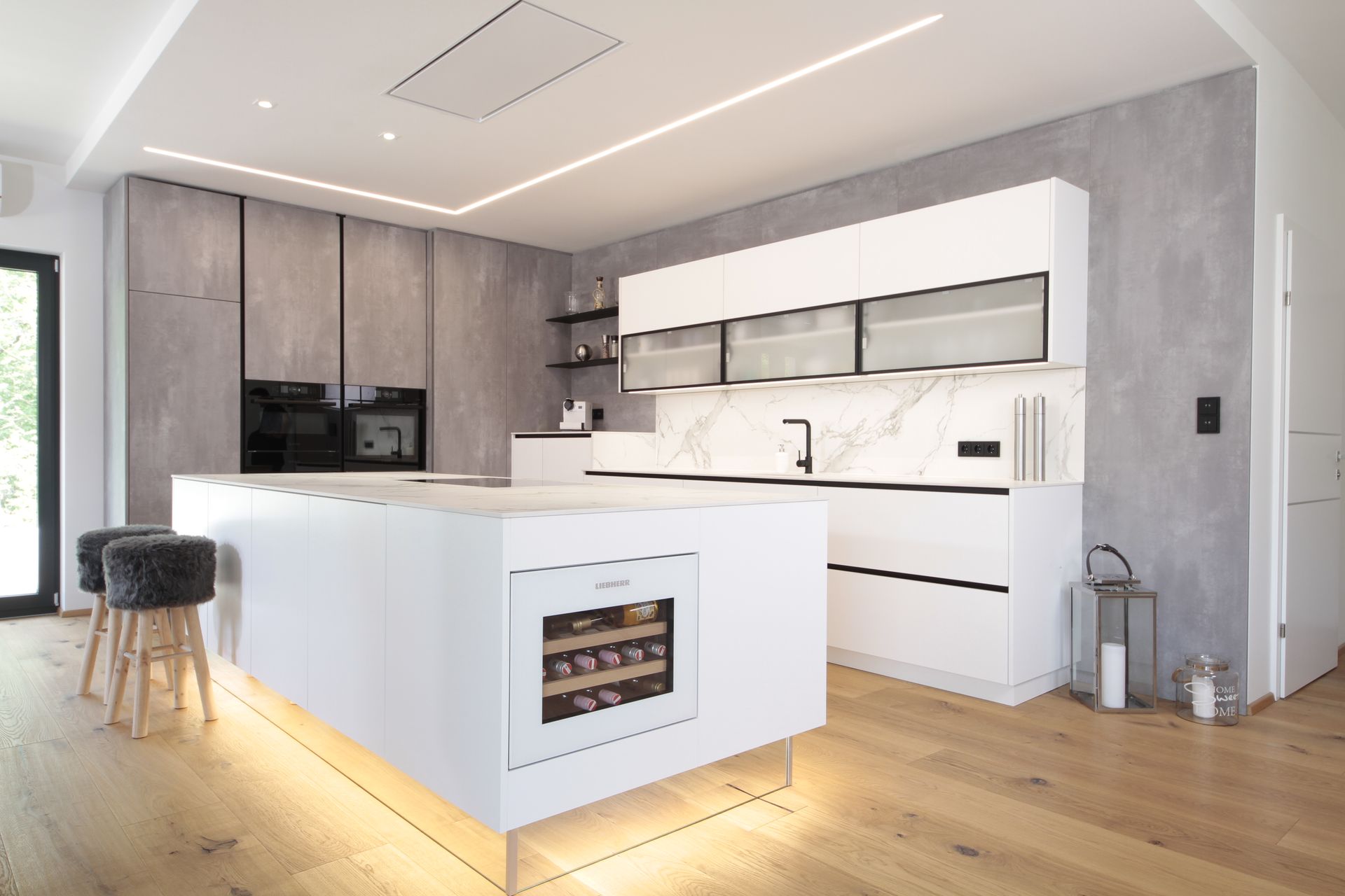 Moderne weiße Küche mit Kochinsel, Einbaubackofen und grauen Holzschränken in einem hellen, minimalistischen Interieur