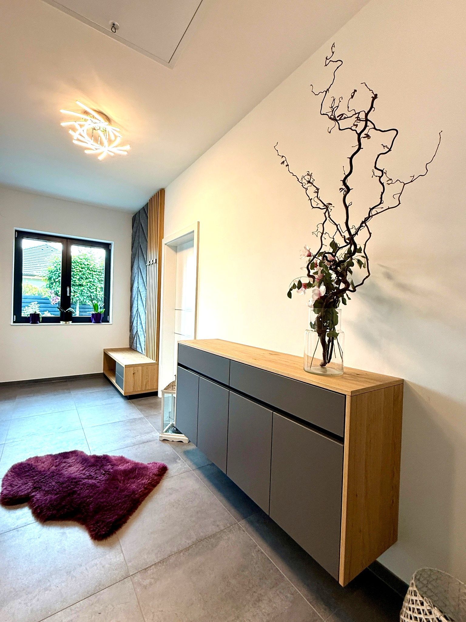 Helles, minimalistisches Zimmer mit niedrigem Schrank, Vase mit Zweigen, Fenster und lila Teppich.