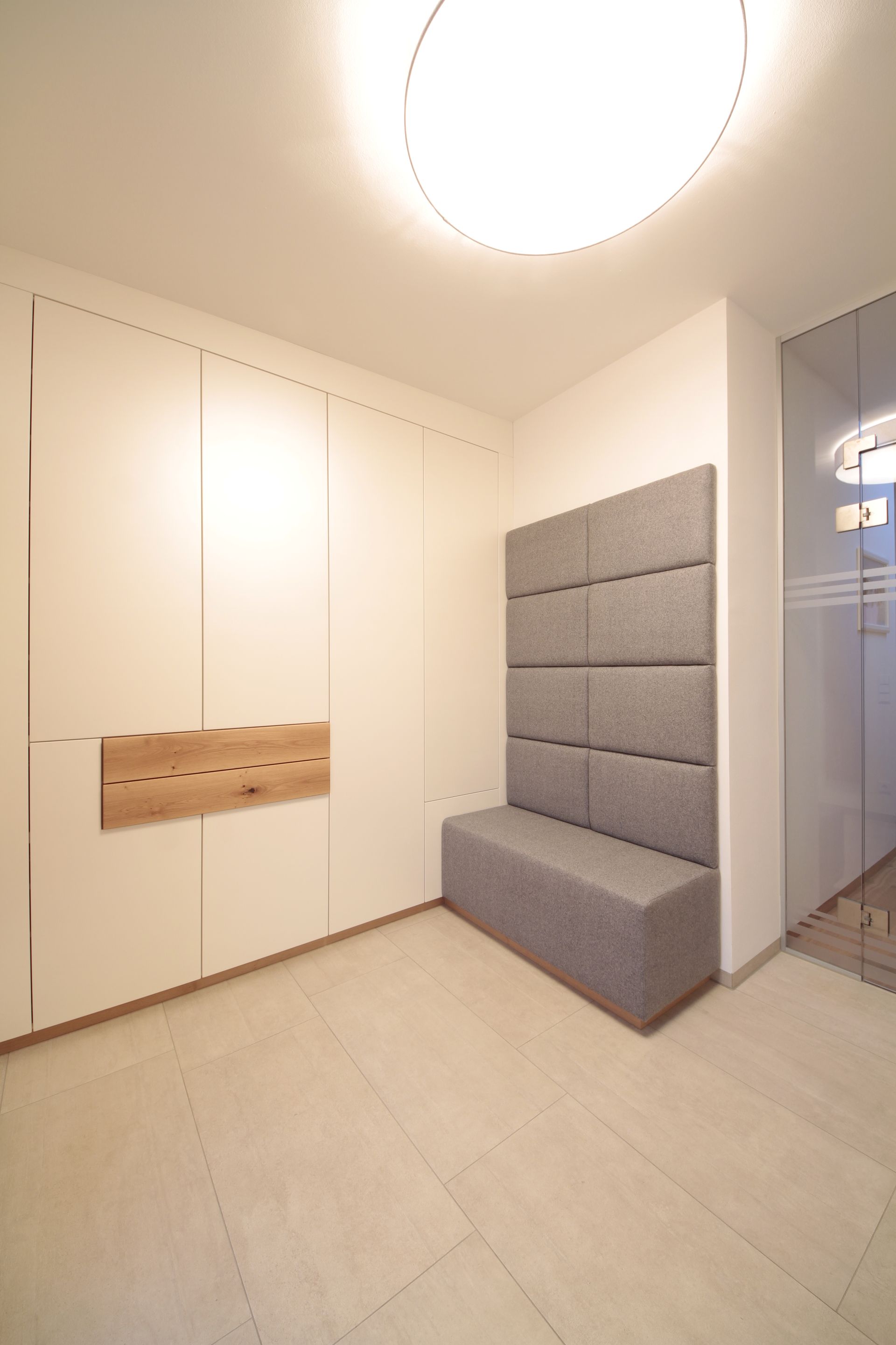 Kleiner Eingangsbereich mit weißen Wänden, einem an der Wand befestigten Holzregal und einer grauen gepolsterten Bank 