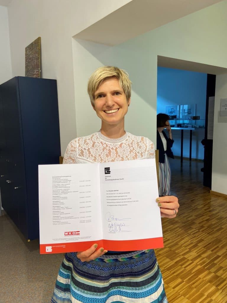 Eine Frau hält ein offenes Dokument in einem Flur und lächelt neben einem dunklen Schrank und einem blau beleuchteten Raum.