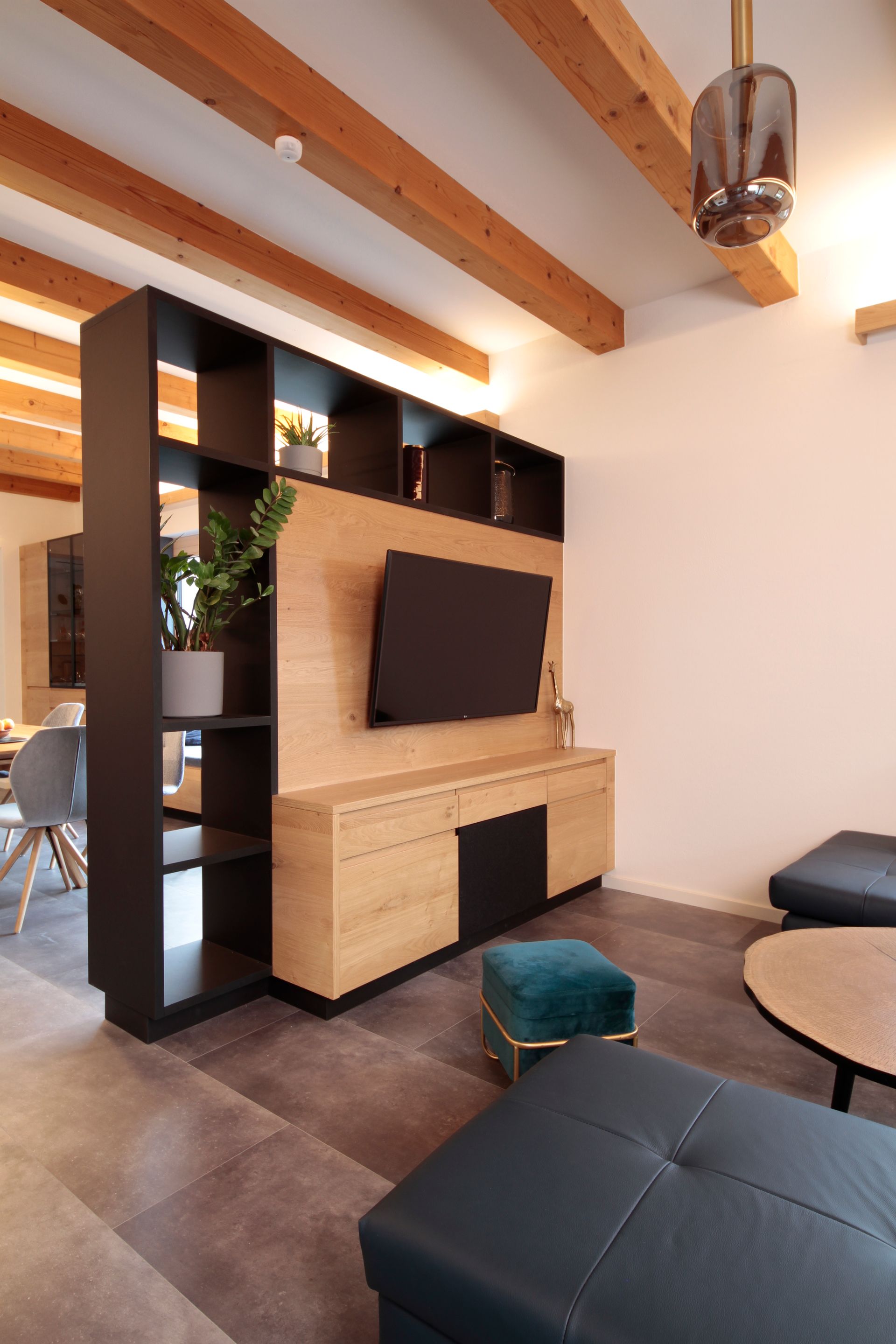 Modernes Wohnzimmer mit Holzbalken, TV-Wandschrank, dunklen Regalen und türkisfarbenem Ottoman.