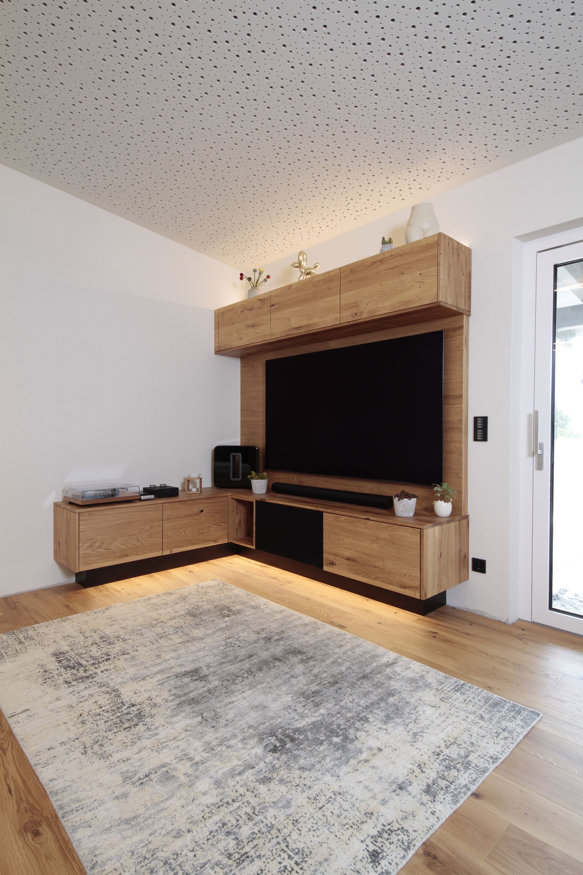 Modernes Wohnzimmer mit großem Fernseher, hölzernem Medienschrank, Beistellschrank, Teppich und warmer Akzentbeleuchtung.