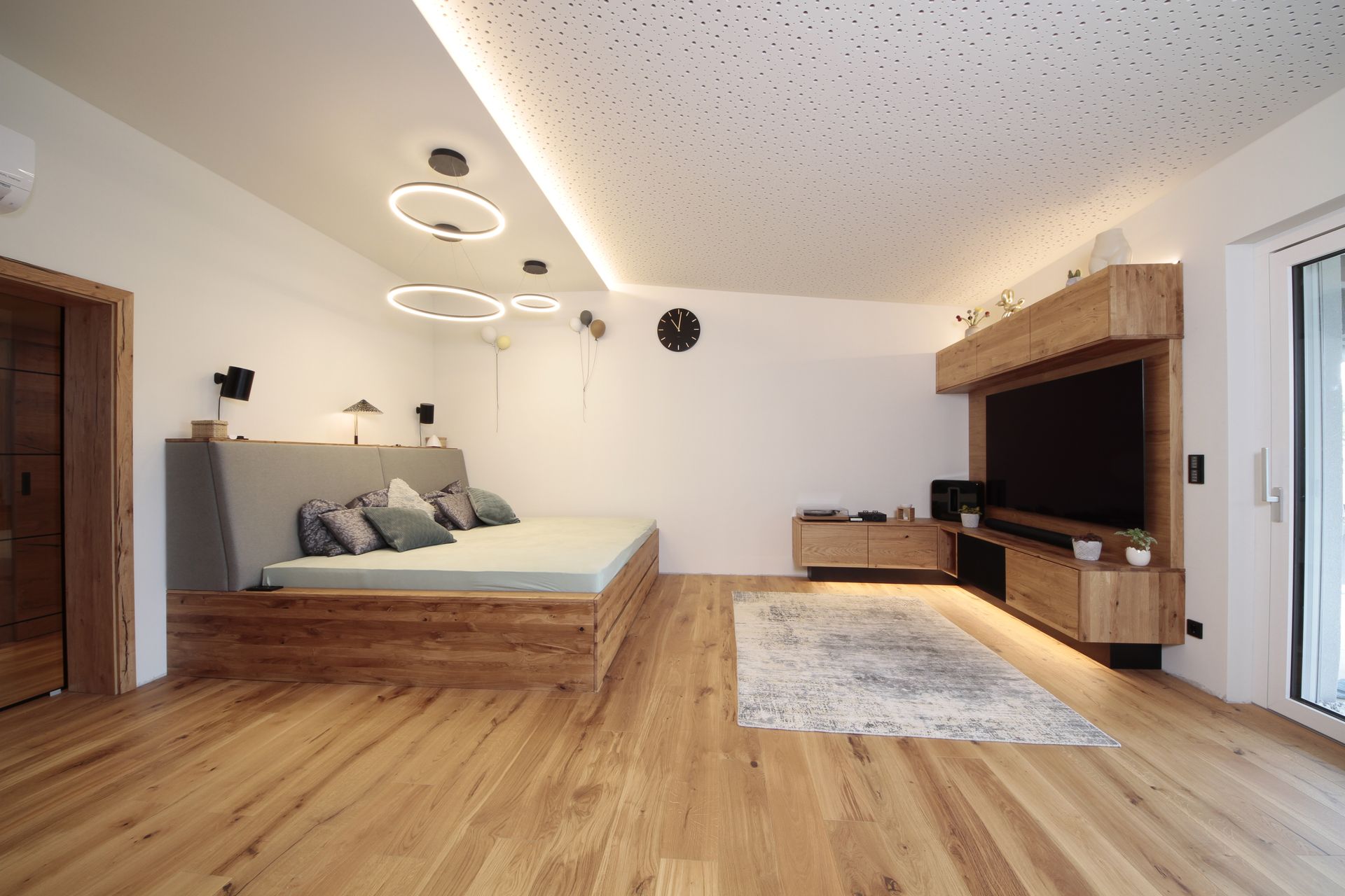 Modernes Schlafzimmer mit grauem Bett, Holzboden, Deckenleuchten und einem TV-Möbel am Fenster.