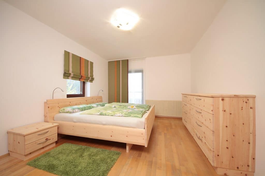 Helles Schlafzimmer mit Bett, Holzkommode, Nachttisch, grünem Teppich und Deckenleuchte im Oberlichtstil.