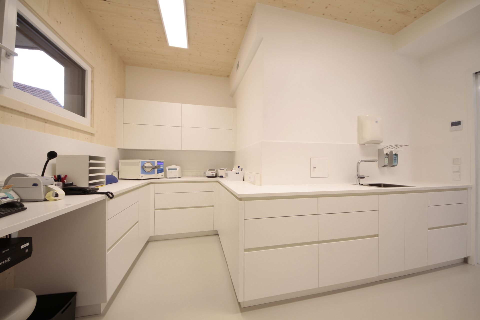 Helles, weißes Untersuchungszimmer mit Schränken, Arbeitsflächen, Waschbecken und Leuchtstoffröhren an der Decke.
