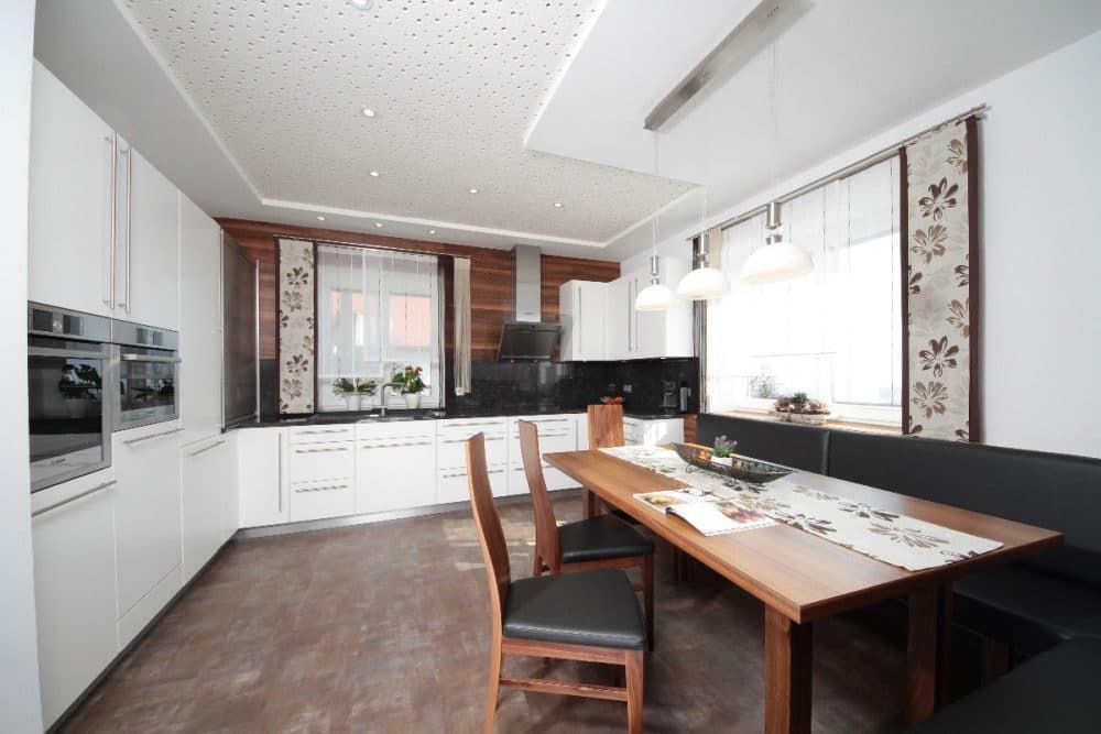 Moderne Küche mit weißen Schränken, dunklen Arbeitsplatten, Esstisch und einem großen Fenster mit Jalousien.
