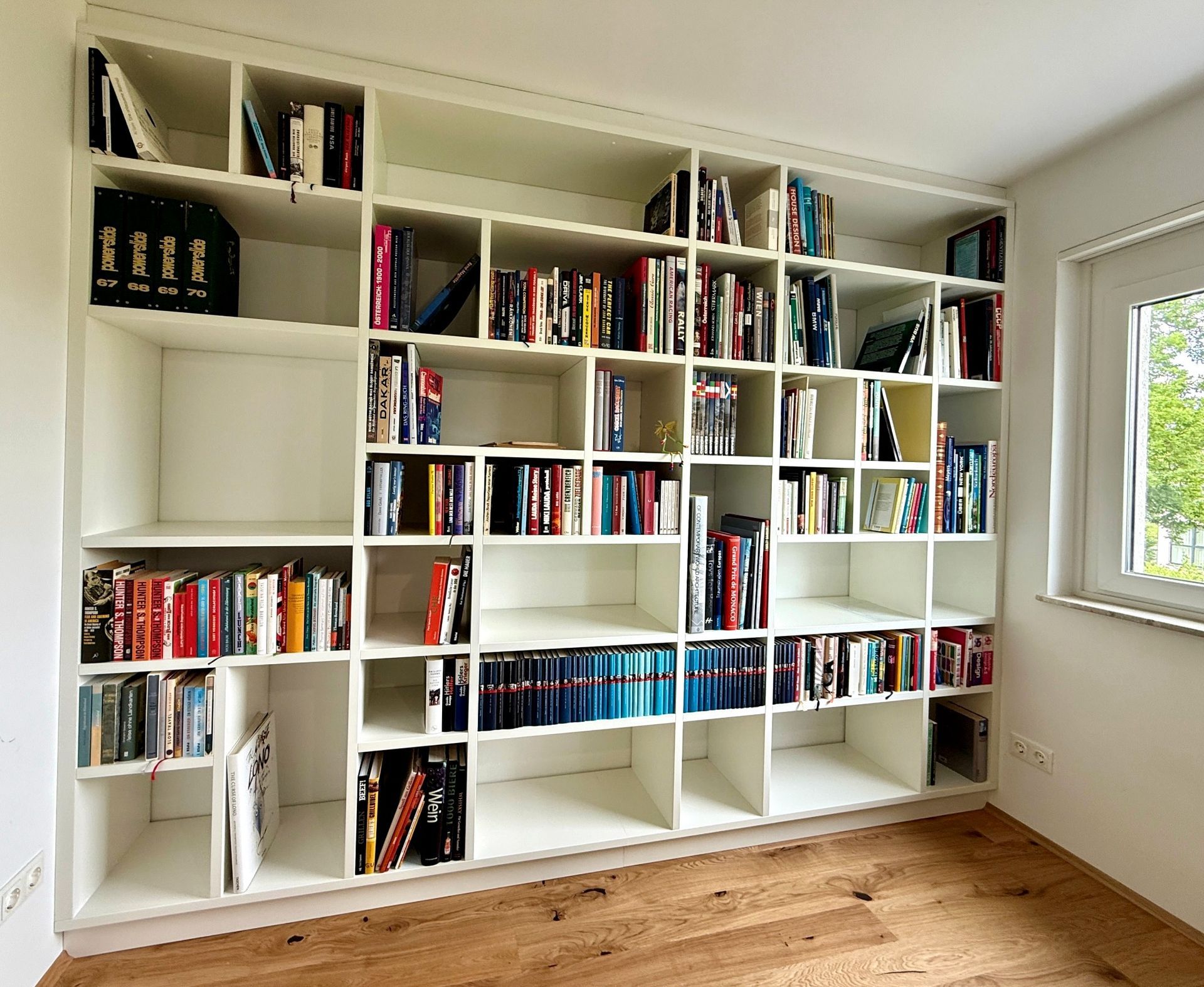 Ein eingebautes weißes Bücherregal voller Bücher in einem hellen Zimmer mit Holzboden und Fenster.