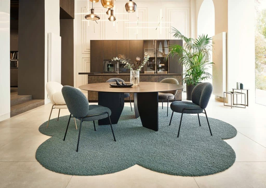 Modernes Esszimmer mit rundem Tisch, vier Stühlen, Kronleuchter und blaugrünem Teppich.