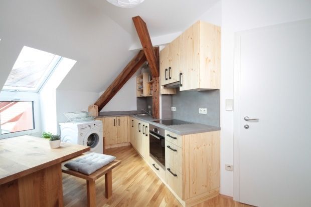 Kleine Dachküche mit Holzschränken, schrägen Deckenbalken, Waschmaschine und Esstisch am Fenster