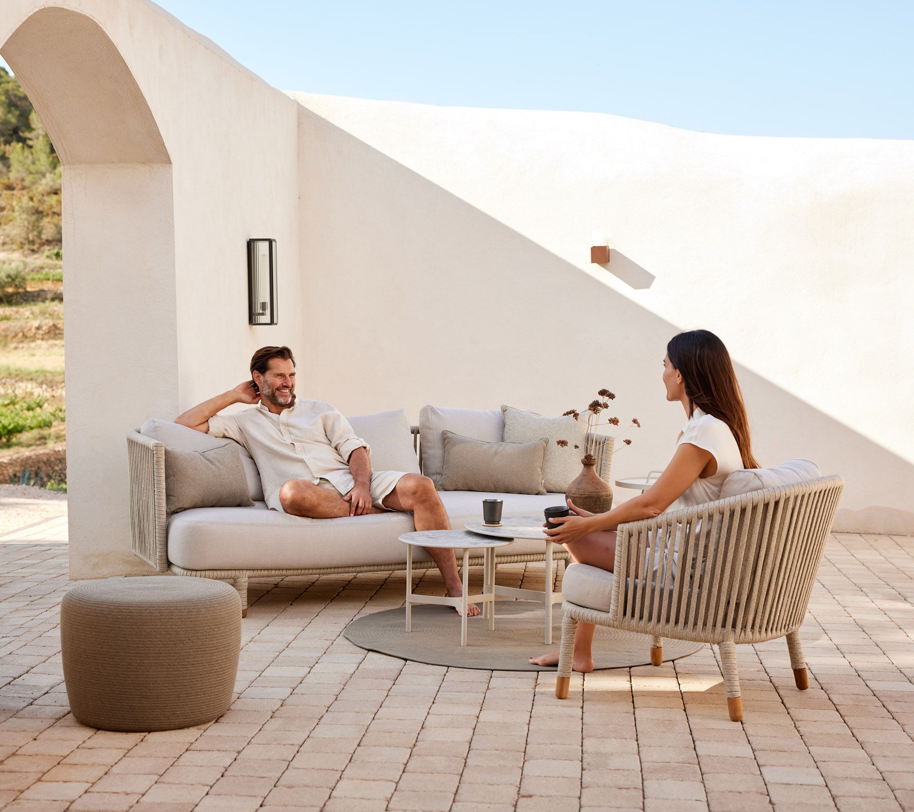 Zwei Personen entspannen sich auf einem beigefarbenen Terrassensofa und einem Stuhl in einem sonnigen Innenhof
