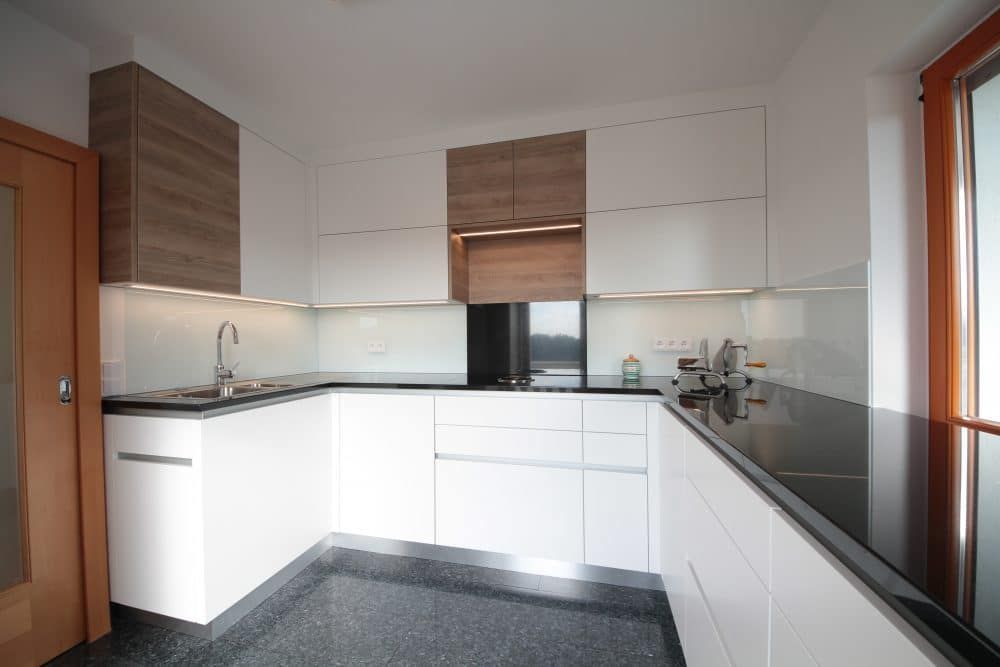 Moderne, weiße L-förmige Küche mit Holzelementen, schwarzen Arbeitsplatten und einem Fenster rechts.