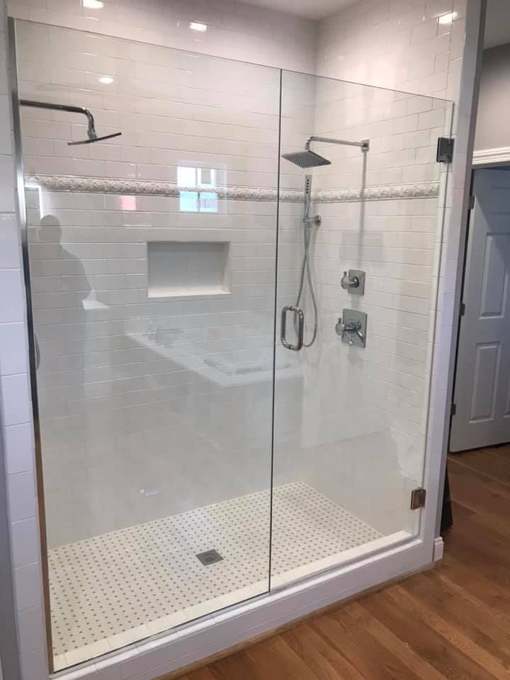 Glass walk-in shower with brass handle in a bright bathroom, next to a window and hanging black robe