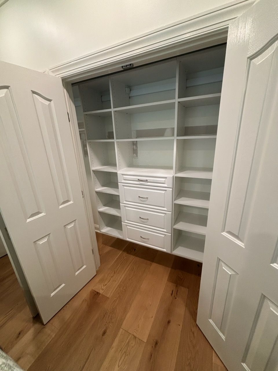Open closet with white built-in shelves and drawers, seen through double doors on a wood floor.