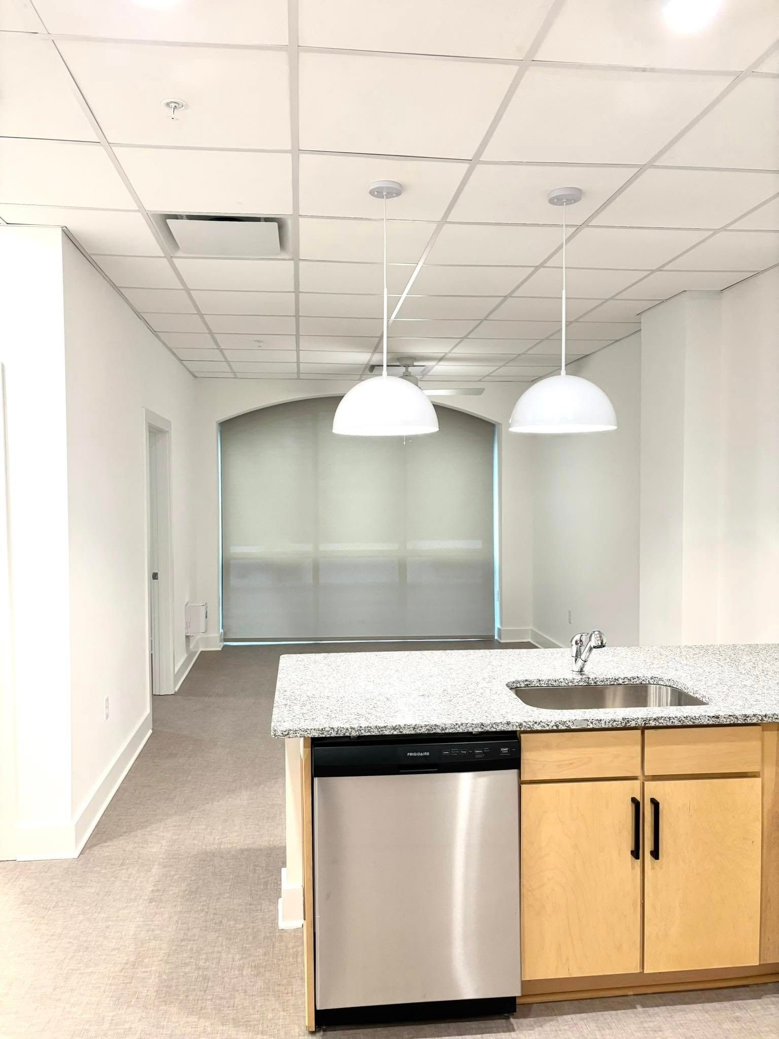 Empty office kitchenette with granite counter, sink, dishwasher, and two pendant lights in a carpeted room