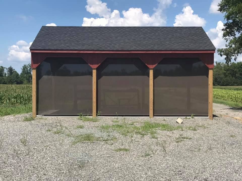 Three-bay wooden shed with dark mesh doors on a gravel lot under a blue sky.