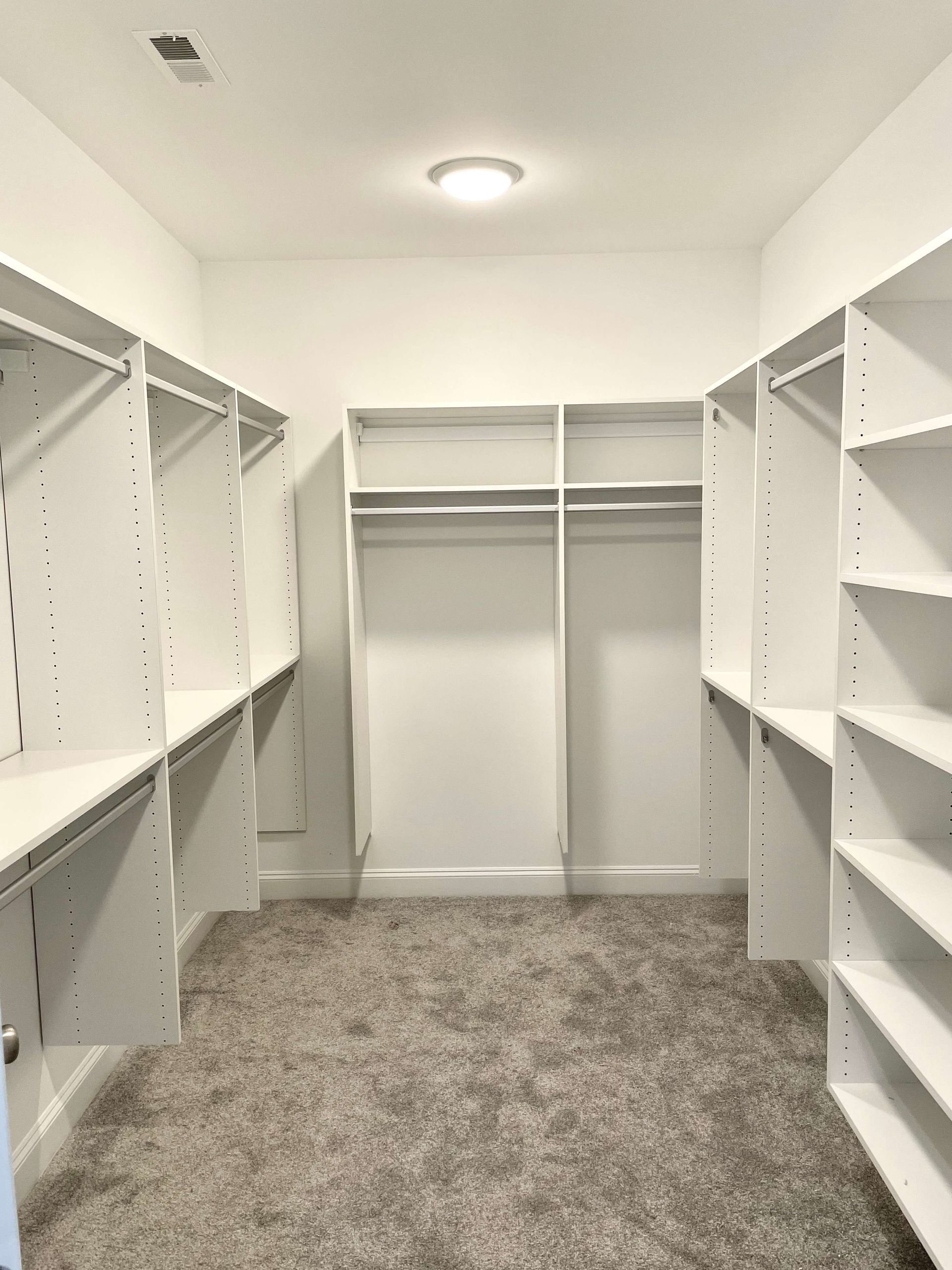 Empty walk-in closet with white shelving and gray carpet, lit by a ceiling light.