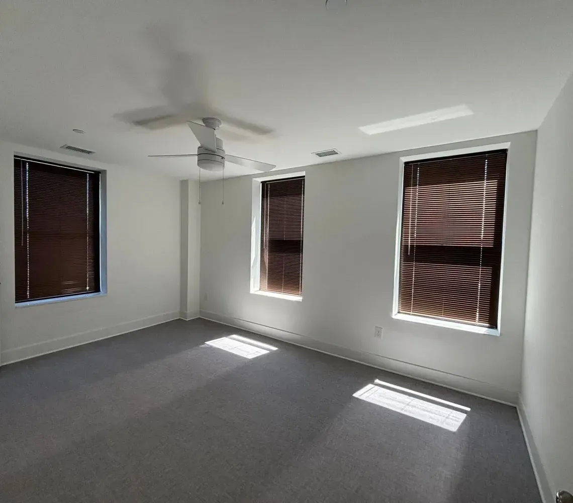 Empty white bedroom with gray carpet, ceiling fan, and three windows with blinds casting sunlight on the floor