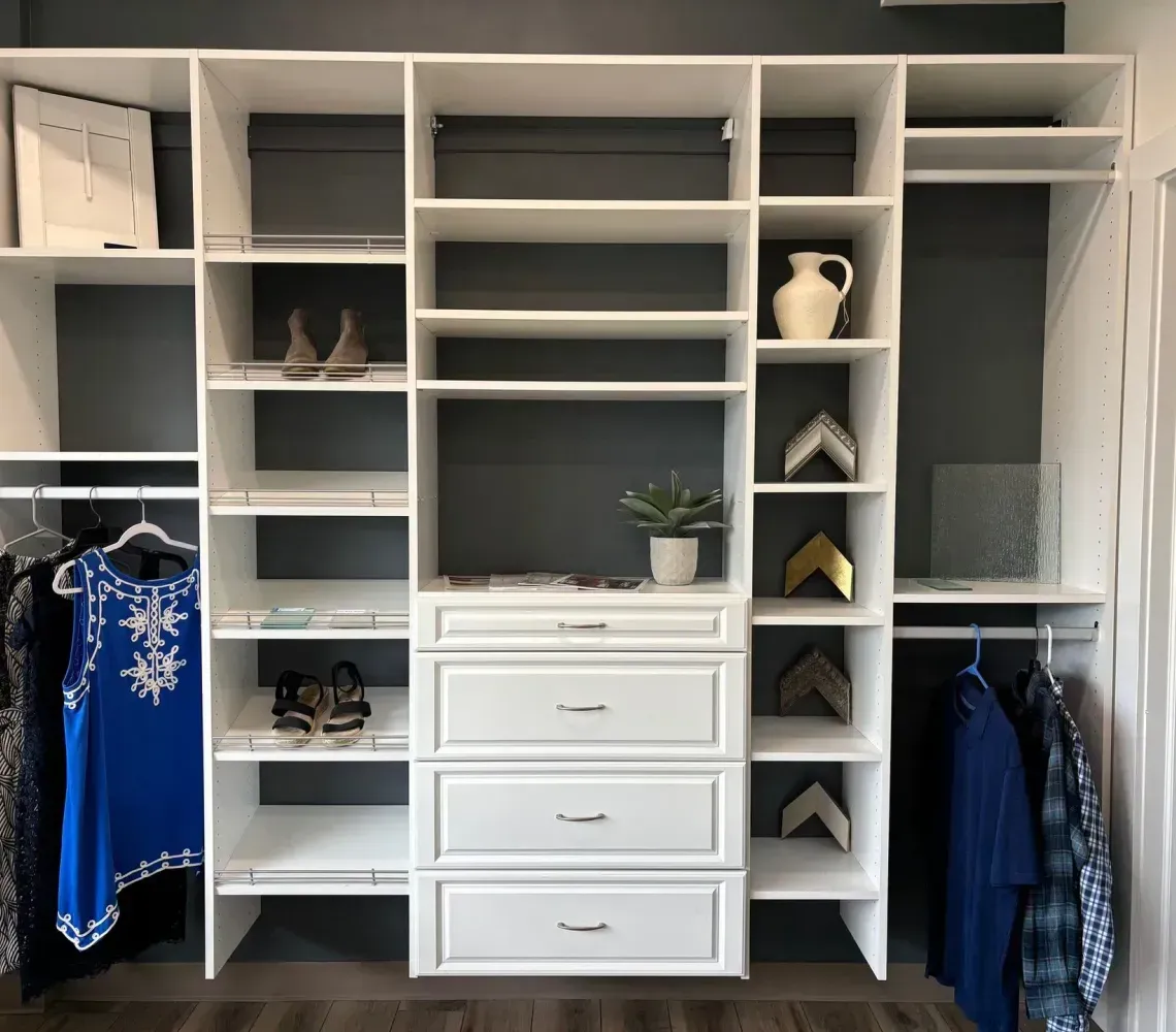 Custom closet with white shelving, drawers, and hanging clothes in a bedroom.