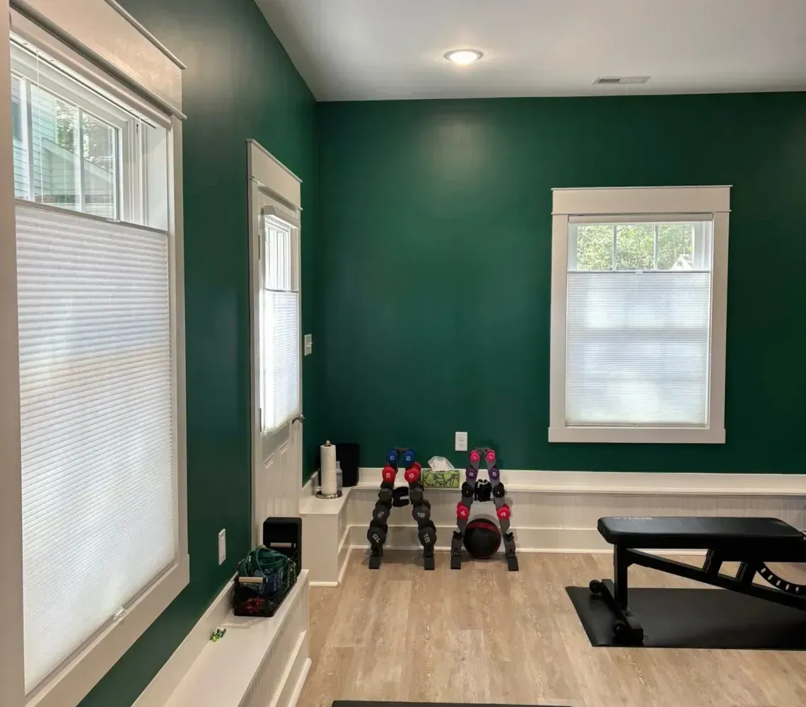 Home gym with green walls, windows, mat, dumbbells, and a small exercise bench on wood floors