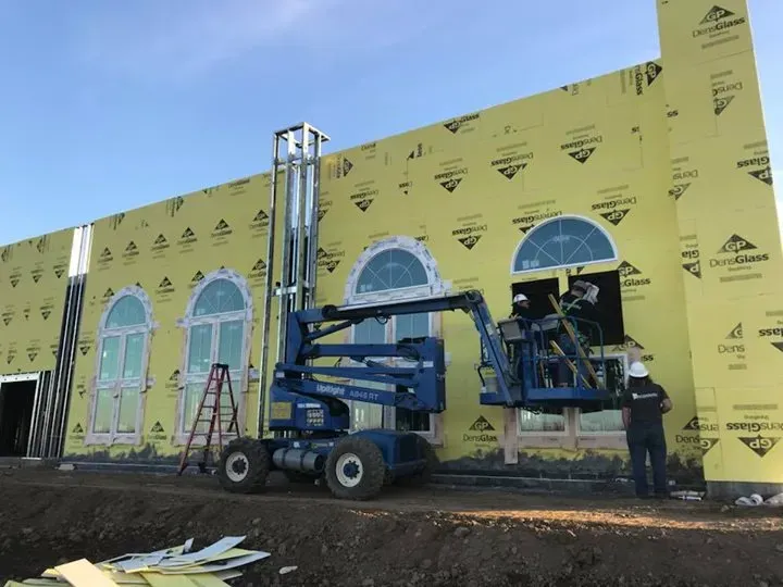 Construction workers install windows on a building exterior using a blue boom lift and a ladder.