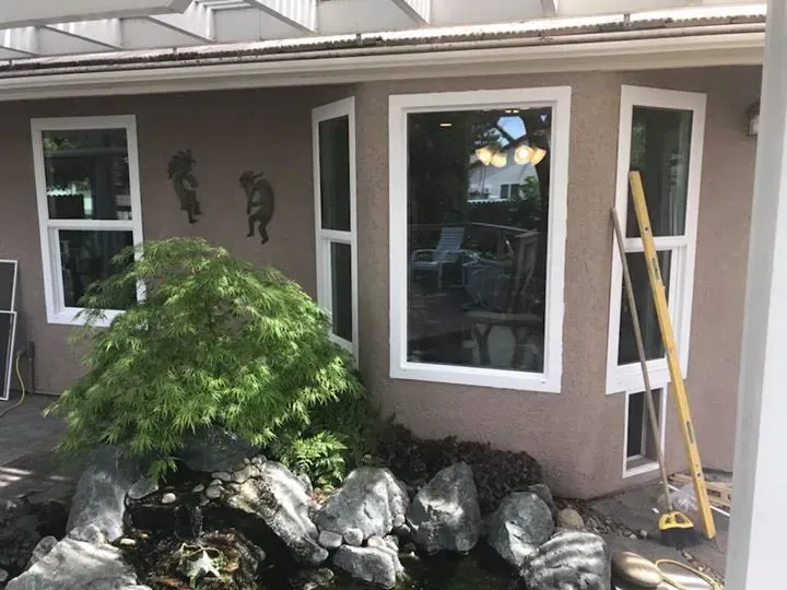 Exterior view of a house with white-framed windows, a small pond with rocks, and green foliage.