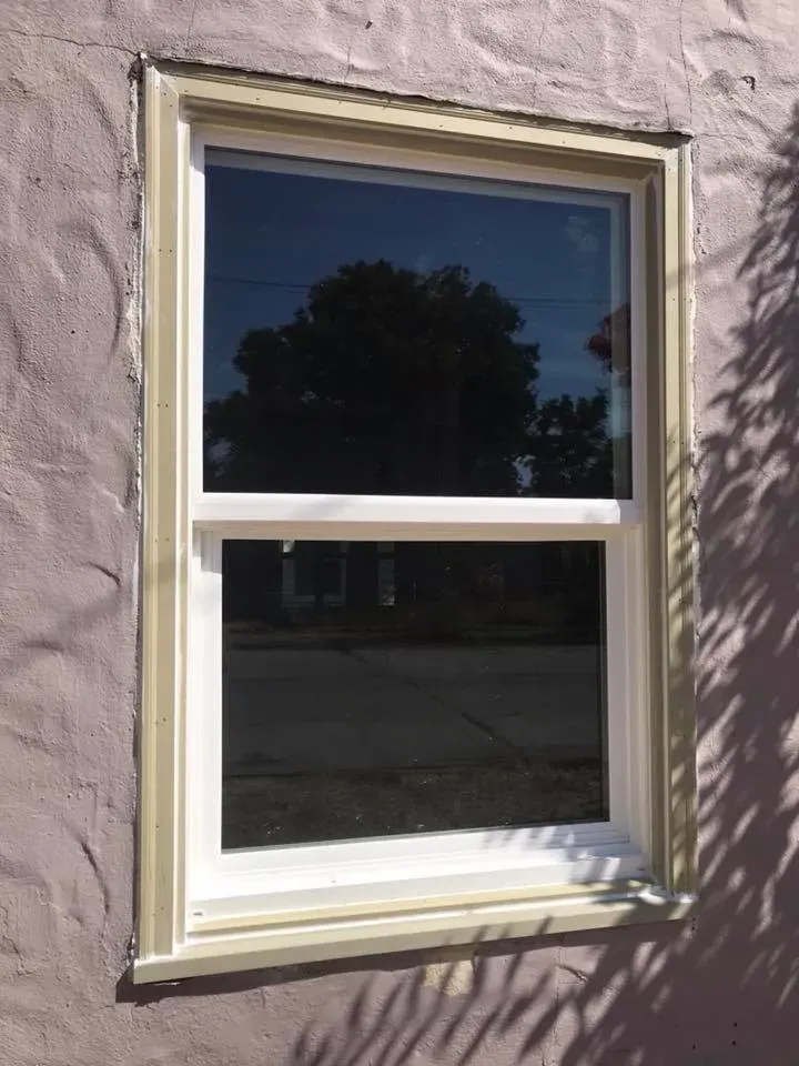 Window with white frame and beige trim in purple stucco wall, reflecting trees and sky.