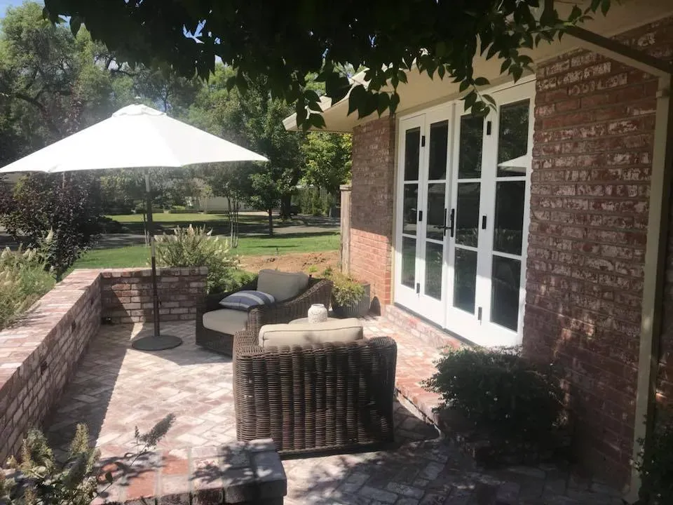 Patio with brick wall and flooring, wicker furniture, and an umbrella, next to a building with glass doors.