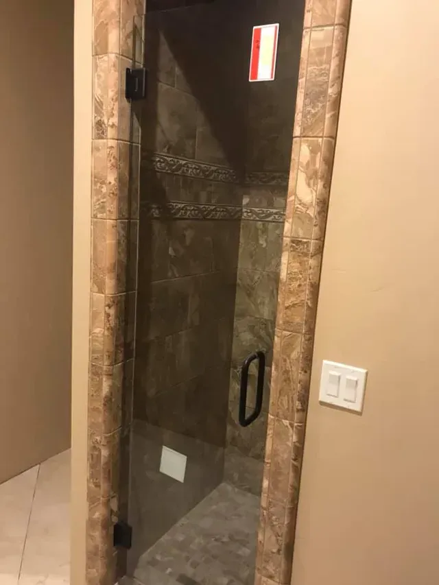 Glass shower door with brown tile frame and handle. Light beige walls surround the door.