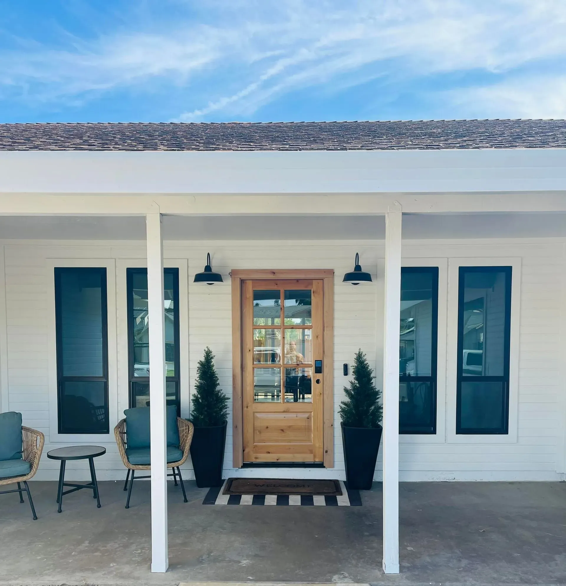 White house front with a wood door, black-framed windows, porch with seating, and potted trees.