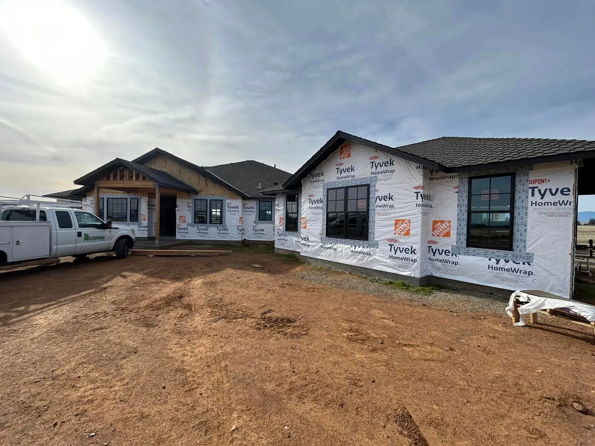 House under construction with tarp-covered walls, black windows, and a partially finished roof on a dirt lot.