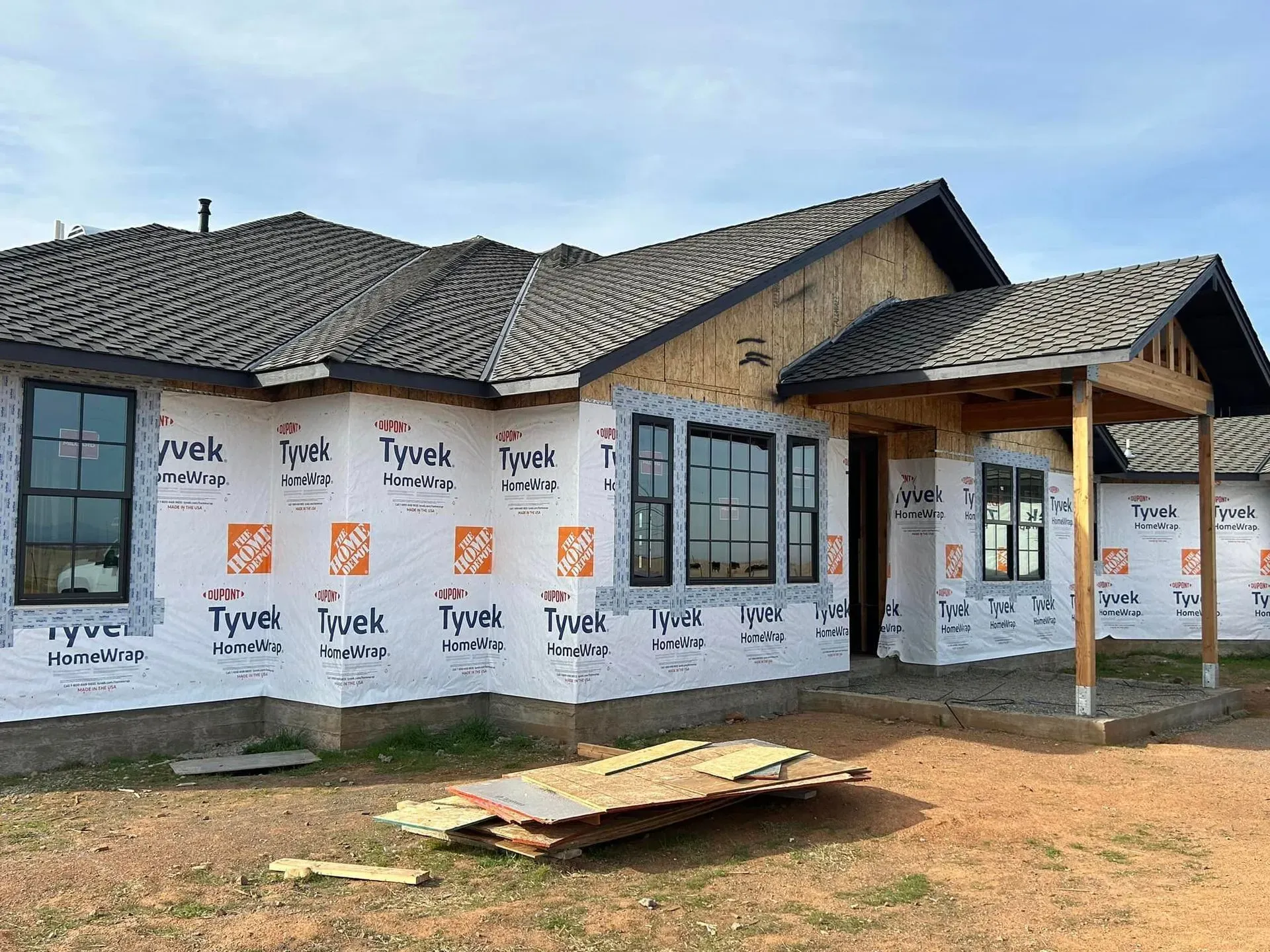 House under construction, covered in Tyvek wrap, with a dark roof and black window frames.