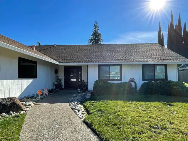 White house with dark trim and roof, sunny day. Walkway leads to front door, greenery and rock border.