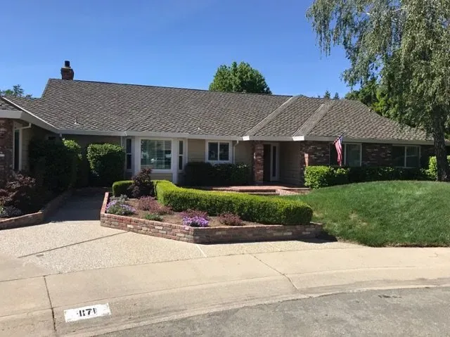 Single-story house with a brick facade, manicured front yard, and a long driveway under a blue sky.