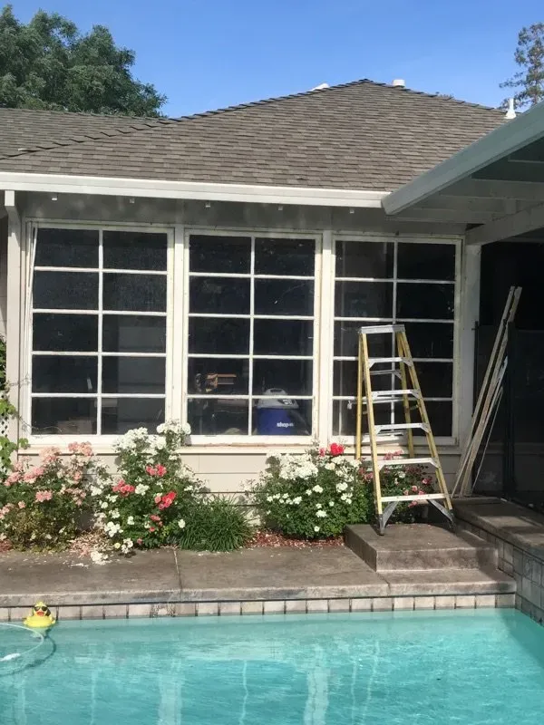 Three-pane window with a ladder, behind a pool. The house has flowers and a brown roof.