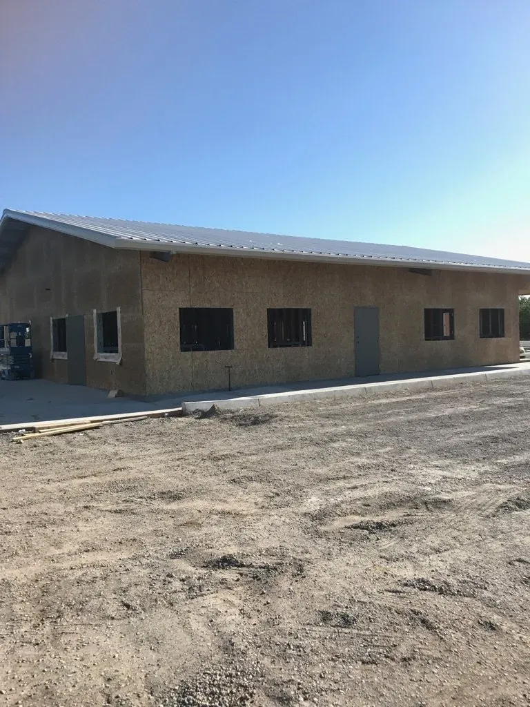 Building under construction, exterior view. Tan wood siding, metal roof, windows and door, dirt ground, clear sky.