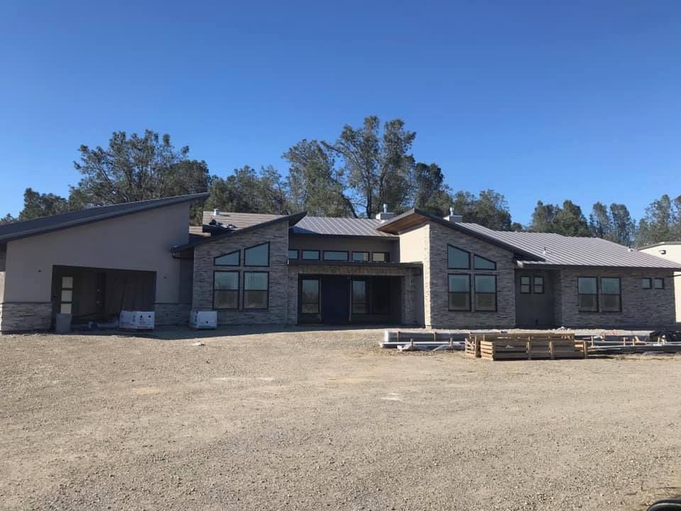 Modern, single-story house under construction with a gray brick facade, large windows, and a metal roof.