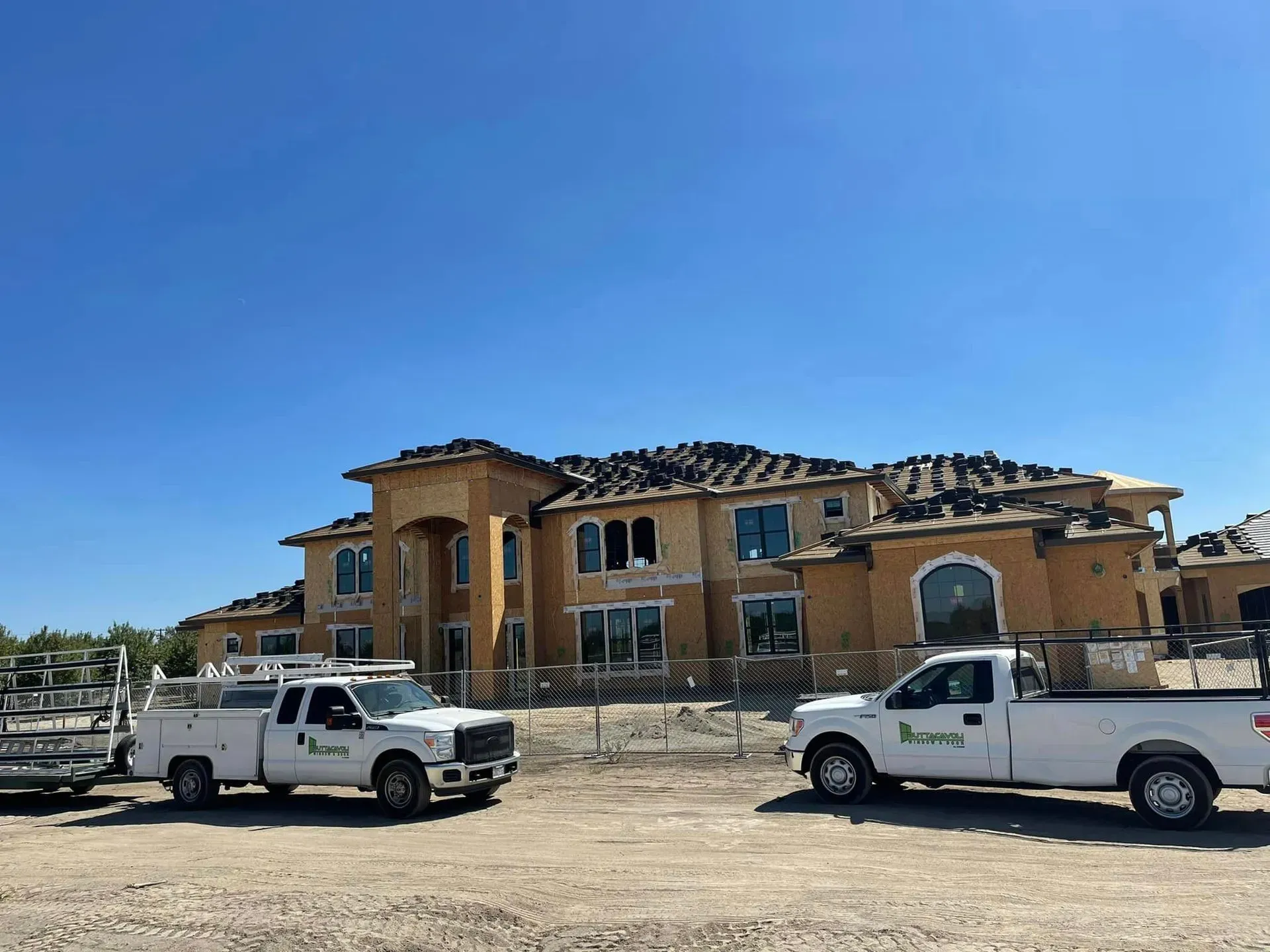 Construction site: Two white work trucks parked in front of a large house under construction; blue sky.