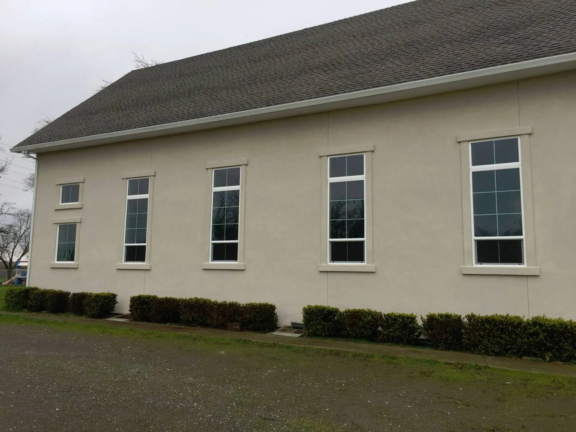 Beige building with tall windows and a dark roof. Low green shrubs line the base.