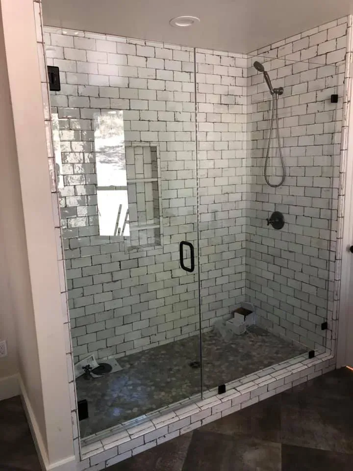Glass shower enclosure with white brick-like tile walls and a gray stone floor, dark hardware.