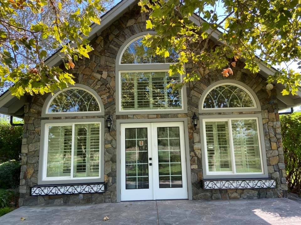 Stone building with arched windows and double doors, surrounded by greenery.