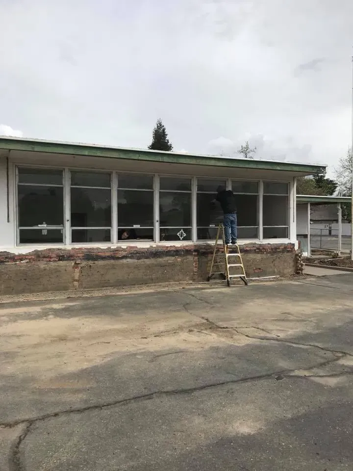 Man on ladder cleaning windows of a low, white building with a brick base on a cloudy day.