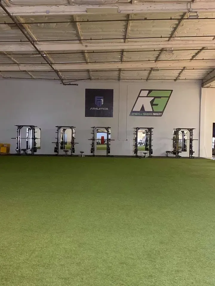 A gym with turf flooring and multiple weight machines. Logos are visible on the wall.