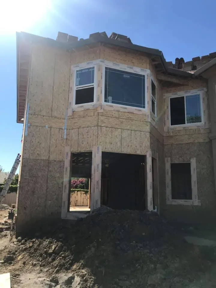 Two-story house under construction, exterior view with sheathing and window frames visible, on a sunny day.