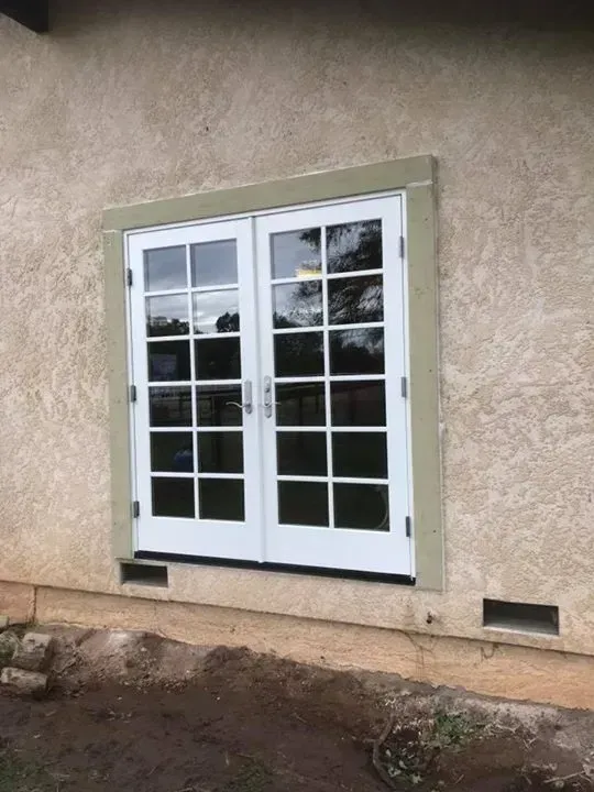 White French doors set into a stucco wall with a green trim. A foundation with vents is visible.
